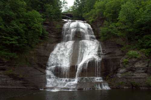 A cascading waterfall flows down rocky cliffs, surrounded by lush green trees and a serene pool at the base.