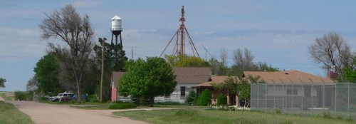 A small rural town with a water tower, a tall structure, and several houses along a dirt road surrounded by greenery.