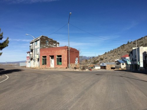 Abandoned buildings at a street corner in a deserted town, with mountains and a clear blue sky in the background.