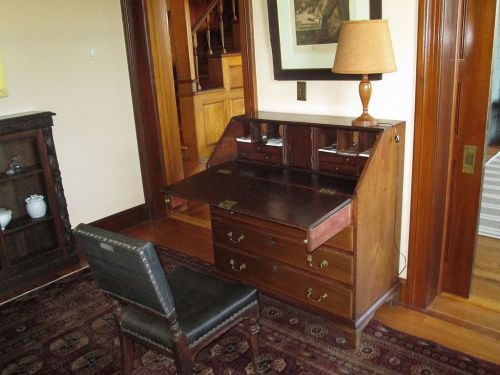 A wooden writing desk with a chair, featuring a pull-down top and drawers, set in a cozy room with warm lighting.