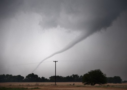 A dark, ominous sky with a funnel cloud forming, set against a rural landscape with a power line and trees.