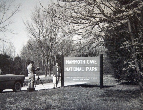 A man films a woman standing by the Mammoth Cave National Park sign, with trees and a vintage car in the background.