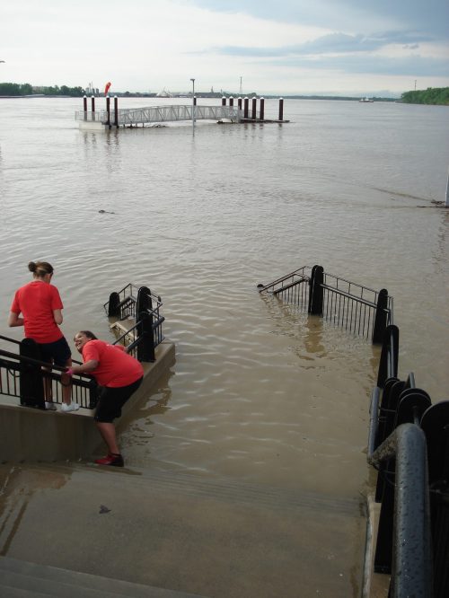 Two people in red shirts are working near a flooded area, with water covering the ground and structures in the background.