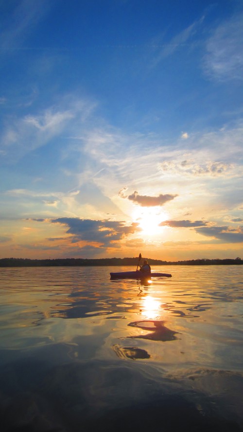 A person kayaking on calm water at sunset, with vibrant clouds and reflections in the water.
