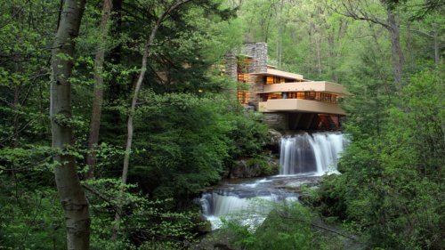 A modern house nestled in lush greenery beside a waterfall, surrounded by trees in a serene natural setting.