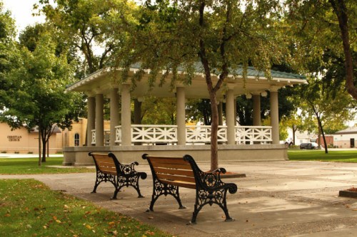 A park scene featuring a gazebo surrounded by trees and two benches on a paved area.