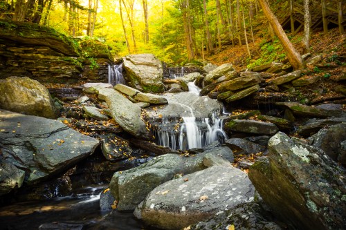A serene waterfall cascades over large rocks, surrounded by vibrant autumn foliage in a peaceful forest setting.