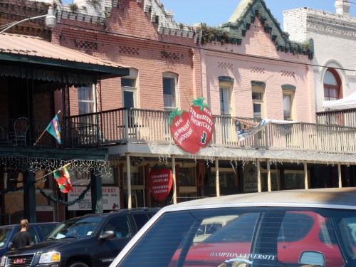 Historic buildings with colorful facades and a large strawberry sign, featuring shops and parked cars in the foreground.