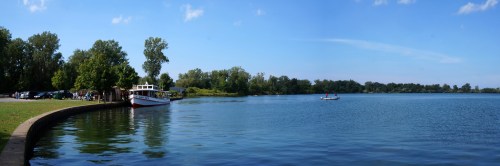 A serene lakeside view with trees, a boat docked, and a calm blue sky reflecting on the water.