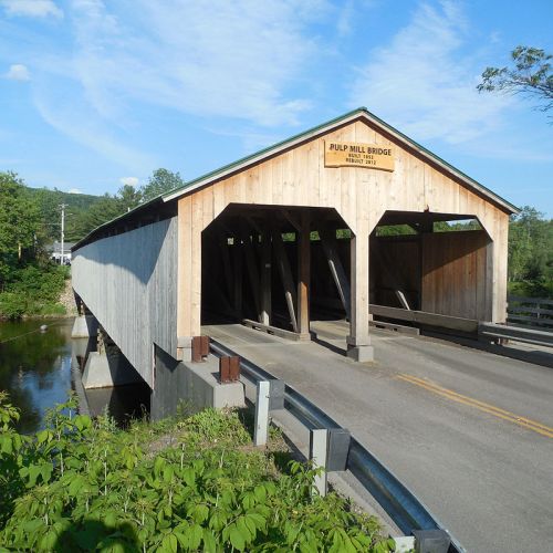 A wooden covered bridge spans a river, with a sign reading "Pulp Mill Bridge" above the entrance. Lush greenery surrounds it.