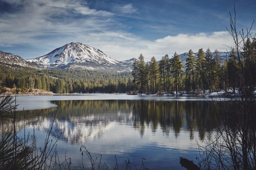 A serene lake reflects a snow-capped mountain and pine trees under a clear blue sky.