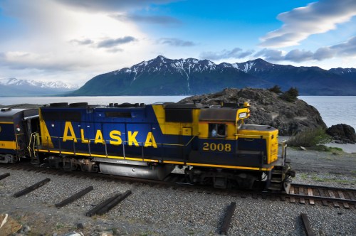 A blue and yellow train labeled "Alaska 2008" travels along a scenic route by the water and mountains.