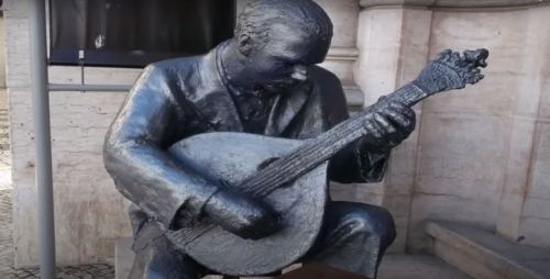 A bronze statue of a man playing a lute, seated outdoors near a building.