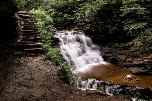 A serene waterfall cascades over rocks, surrounded by lush greenery and a stone staircase nearby.