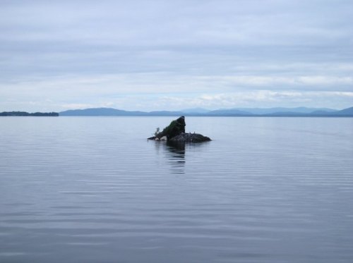 A small rocky island in a calm body of water, surrounded by distant mountains under a cloudy sky.