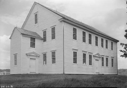 A historic white wooden building with multiple windows, set against a cloudy sky and grassy foreground.