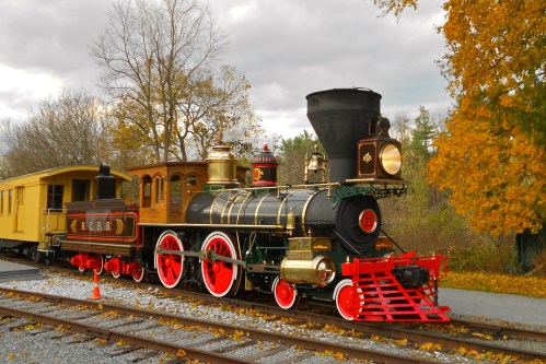 A vintage steam locomotive with a black and gold design, parked on tracks surrounded by autumn trees.