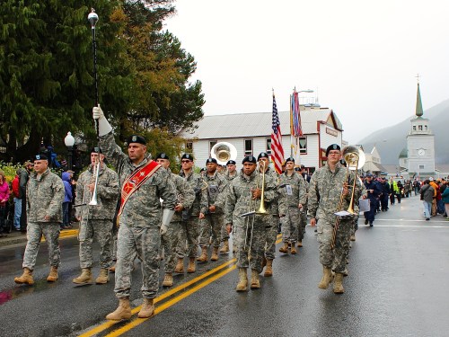 A military band in uniform marches down a rainy street, with flags and a church in the background.