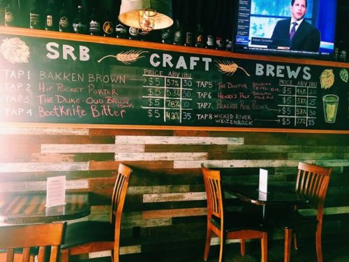 Chalkboard menu displaying craft beers with prices and details, alongside wooden chairs in a cozy bar setting.