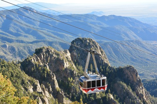 A cable car glides over rugged mountains, surrounded by lush greenery and a vast landscape in the distance.