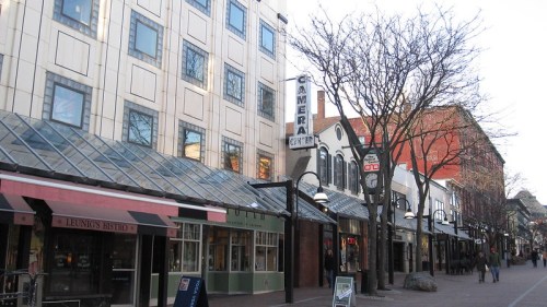 A street view featuring shops and storefronts, with trees and a clear sky in the background.