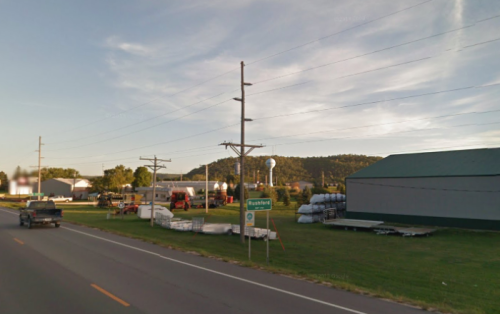 A rural scene featuring a road, utility poles, buildings, and a water tower against a backdrop of hills.
