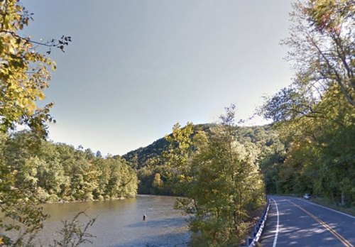 A serene river scene with a fisherman, surrounded by lush trees and a winding road under a clear sky.