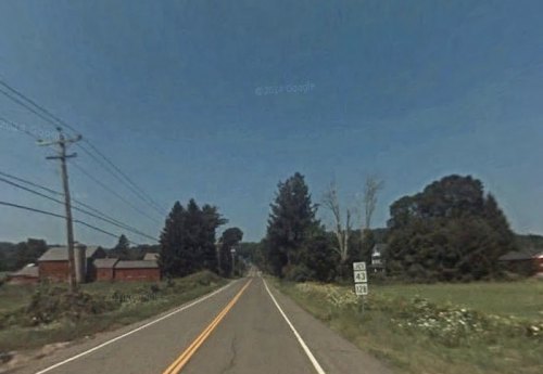 A rural road with power lines, flanked by fields and trees, leading towards distant buildings under a clear blue sky.