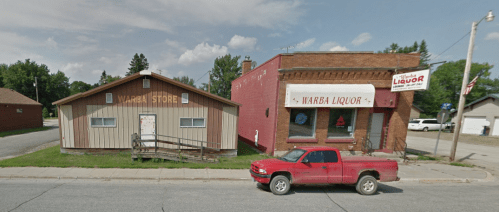 A red pickup truck parked in front of two buildings: a wooden store and a brick liquor store with signs.
