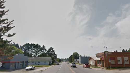 A quiet street scene with buildings on either side, trees in the background, and a cloudy sky overhead.