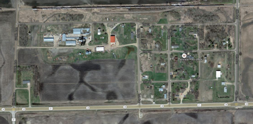 Aerial view of a rural area with fields, buildings, and a small community layout. Roads and structures are visible.