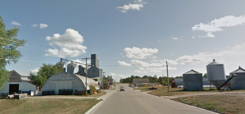 Rural scene featuring a road lined with silos and farm buildings under a blue sky with scattered clouds.