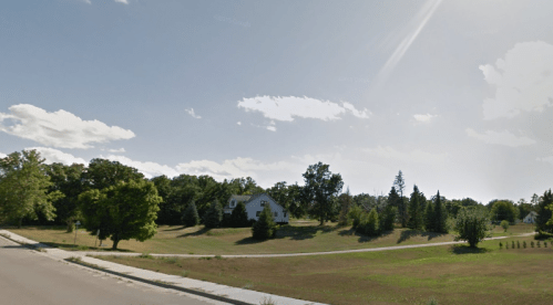 A sunny landscape featuring a grassy area, trees, and a house in the distance under a clear blue sky.