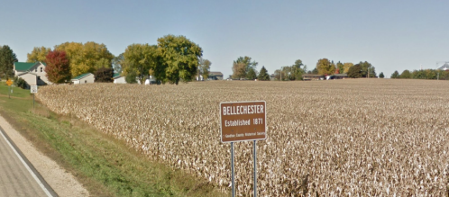 Sign for Bellechester, established 1871, with a cornfield and trees in the background on a clear day.