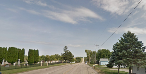 A quiet road lined with trees, leading to a cemetery on one side and a small house on the other under a blue sky.