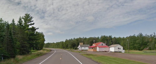 A rural road curves past a few houses and trees under a partly cloudy sky.