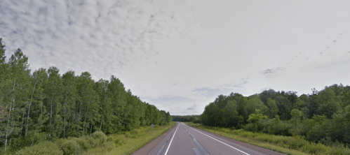 A wide, empty highway stretches through lush green trees under a cloudy sky.