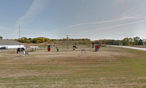 A grassy area with sculptures, including a large red object and a figure, set against a backdrop of trees and blue sky.