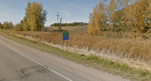 A roadside sign indicating "Visiting" near a field with tall grass and trees in autumn colors.