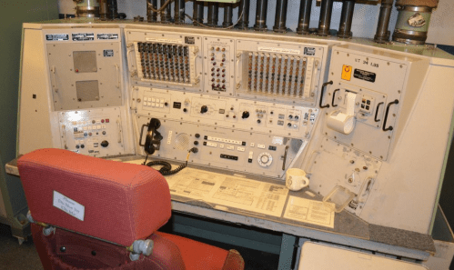 A vintage control panel with buttons, dials, and a red chair, set in a technical environment.