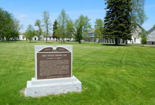 A historical marker stands in a grassy area, with trees and old buildings visible in the background at Fort Totten.