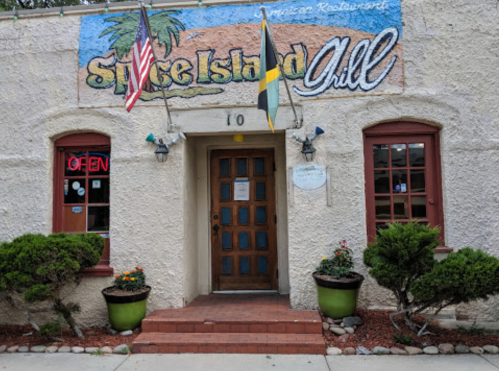 Entrance of Spice Island Chill restaurant with flags, open sign, and potted plants. Colorful mural above the door.