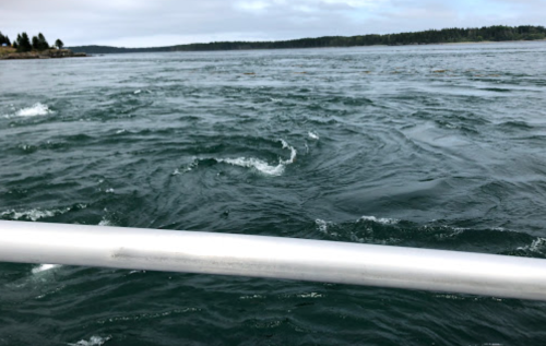 A close-up view of swirling water with a silver railing in the foreground, set against a cloudy sky.