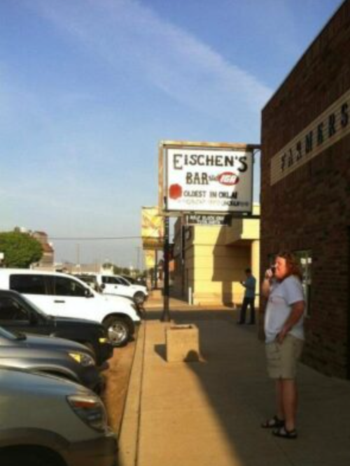 A man stands near a sign for Eischen's Bar, with parked cars and a clear blue sky in the background.