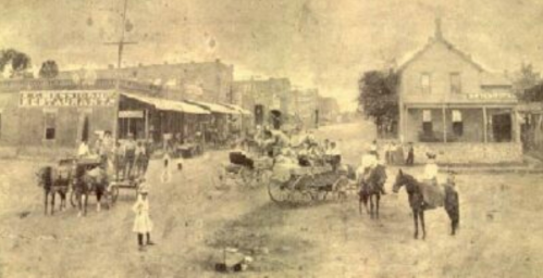 Historic street scene with horse-drawn carts, people, and buildings in a dusty town setting.