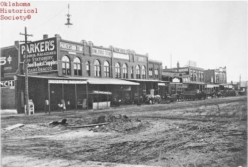 Historic black-and-white photo of a street lined with storefronts and horse-drawn carriages.