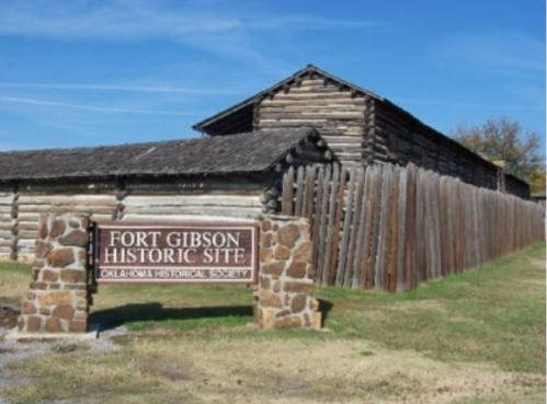 Sign for Fort Gibson Historic Site in front of a log building and wooden fence under a clear blue sky.