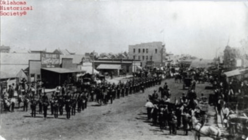 Historic black-and-white photo of a parade in a town, featuring soldiers and crowds in a bustling street scene.