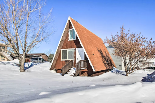 A snow-covered A-frame cabin with a wooden deck, surrounded by trees and a clear blue sky.