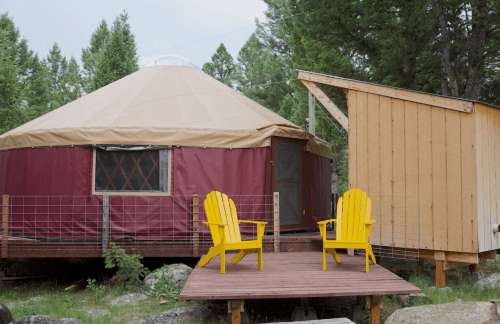 A cozy yurt with a tan roof and maroon walls, accompanied by a wooden deck and two yellow chairs outside.